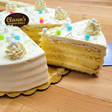 Load image into Gallery viewer, Sliced vanilla custard torte with buttercream frosting and colorful edible pearls on top, on a wooden surface.