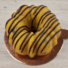 Load image into Gallery viewer, A small yellow cake bundt with dark chocolate drizzle, presented on a round copper-colored plate against a light wood background.