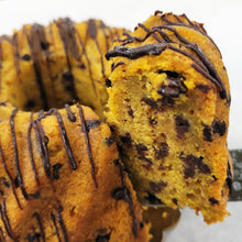 Load image into Gallery viewer, A close-up of a chocolate chip cake with a slice cut out and leaning against the main loaf. The golden-brown baked good is topped with dark chocolate drizzle, and chocolate chips are visible throughout.