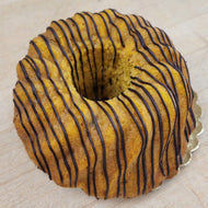 A golden-brown ring cake with dark chocolate drizzle, presented on a light wood surface.