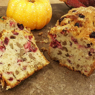 A loaf of cranberry walnut quick bread with a slice cut out to show the filling, placed on a dark surface with a pumpkin and more bread in the background.